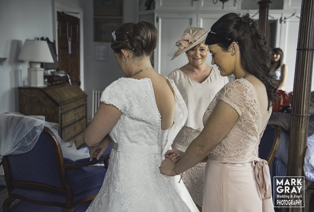 Chief bridesmaid helps bride into dress as mum looks on